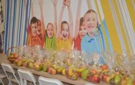 Przygotowane pakiety owocowe dla zaproszonych gości. W tle kolorowa fototapeta z dziećmi.