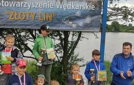 Nagrodzona grupa chłopc&oacute;w w danej kategorii pozuje trzymając trofea na tle baneru stowarzyszenia. Obok stoi prezes stowarzyszenia. W tle jezioro.