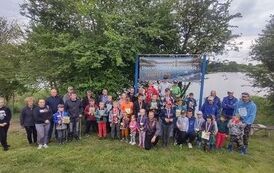 Uczestnicy zawod&oacute;w pozuja trzymając w rękach trofea z opiekunami i sędziami na tle baneru stowarzyszenia. W tle jezioro.