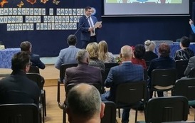 Widok na salę od tyłu. Na pierwszym planie goście siedzący na krzesłach. Na scenie przemawia przewodniczący rady.