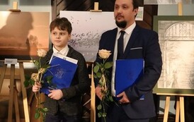 Wsp&oacute;lne zdjęcie tegorocznych stypendyst&oacute;w. Każdy trzyma r&oacute;że i teczkę. W tle prace plastyczne.