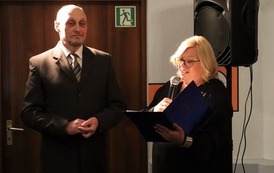 Prowadzący Galę. Na stojąco. Kobieta w jeden ręce trzyma i mikrofon, do kt&oacute;rego m&oacute;wi. W drugiej ręce trzyma otwartą teczkę.