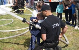 Strażak i dw&oacute;jka dzieci trzymają wąż strażacki, z kt&oacute;rego leci woda.