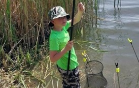Młody wędkarz pozuje do zdjęcia trzymając wędkę i stojąc na brzegu jeziora, na swoim stanowisku.