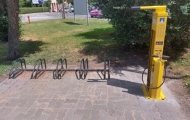 Stacja napraw rower&oacute;w w kolorze ż&oacute;łtym posadowiona na utwardzonym terenie. Obok stojak na rowery.