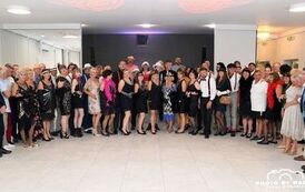 W sali restauracyjnej, w p&oacute;łokręgu pozują uczestnicy balu w strojach z epoki.