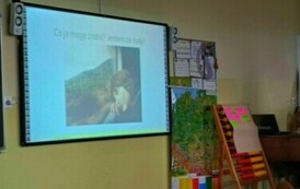 W klasie. Na ścianie z lewej strony wisi ekran, na kt&oacute;rym jest prezentacja. Z prawej - mapa i duże liczydło.