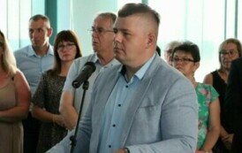 Na pierwszym planie mężczyzna w garniturze stoi przed mikrofonem. W tle stoi kilka innych os&oacute;b.