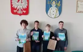 Salka urzędu. Na tle herbu Trzemeszna i godła do zdjęcia pozuje grupa uczni&oacute;w. Każdy trzyma dyplom i papierowa torbę z upominkami.