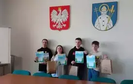 Salka urzędu. Na tle herbu Trzemeszna i godła do zdjęcia pozuje grupa uczni&oacute;w. Każdy trzyma dyplom i papierowa torbę z upominkami.