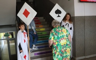 Przy wejściu na schody stoi dw&oacute;jka dzieci ubranych w białe peleryny z karcianymi symbolami. W rękach trzymają kije z przyczepionymi karcianymi znakami.