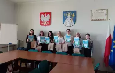 Salka urzędu. Na tle herbu Trzemeszna i godła do zdjęcia pozuje grupa uczni&oacute;w. Każdy trzyma dyplom i papierowa torbę z upominkami.