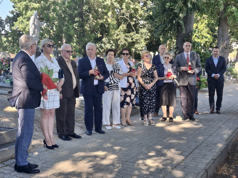 Członkowie komitetu stoją na chodniku w alejce cmentarnej. Niektórzy trzymają znicze.